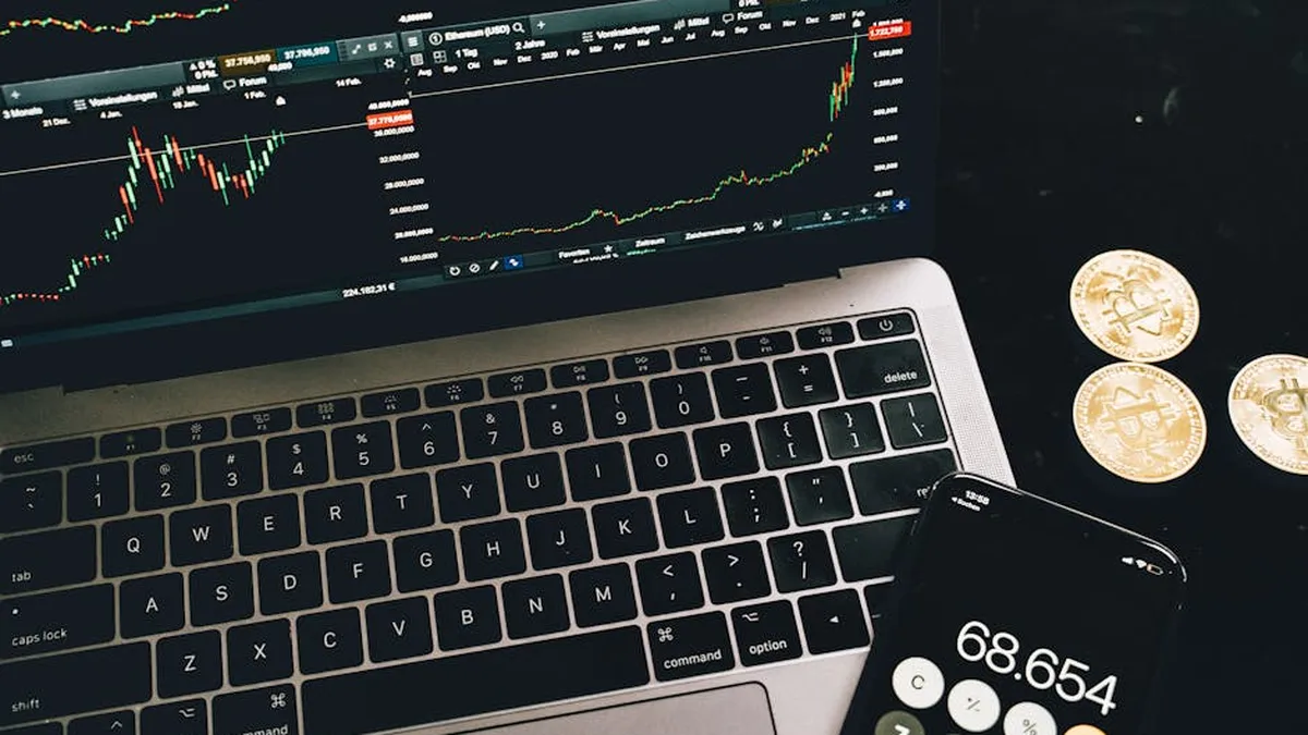A laptop with trading charts, smartphone calculator, and bitcoin coins depicting cryptocurrency trading.