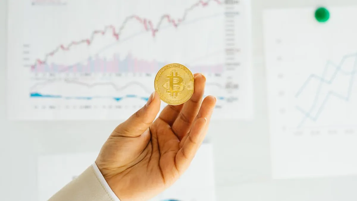A hand holding a Bitcoin coin against financial charts depicting market trends.