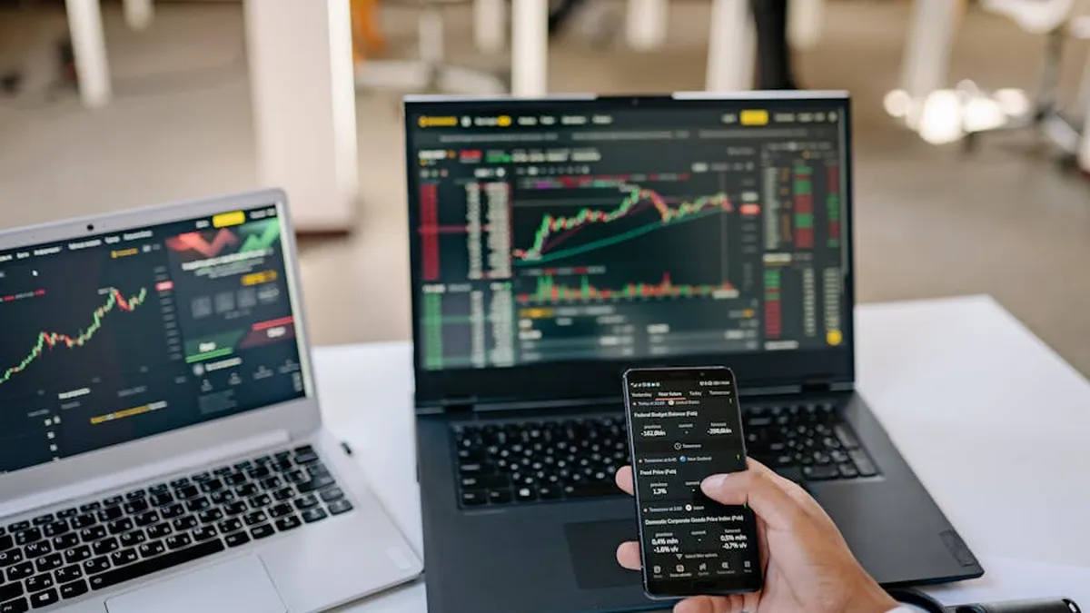 A professional tracks stock market trends using two laptops and a smartphone, showcasing digital finance tools.