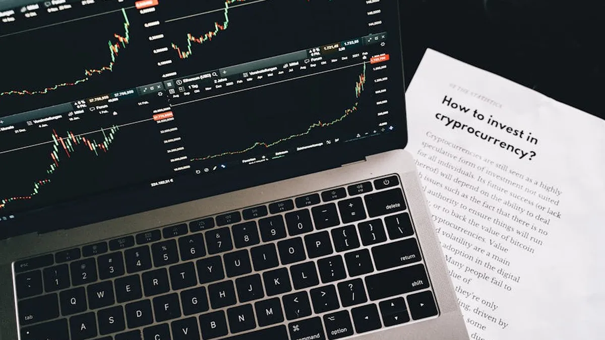 Close-up of a laptop displaying stock market graphs with a paper on investing in cryptocurrency, indoors.