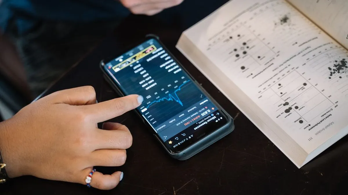 Hand pointing at smartphone displaying financial data, with open book nearby.