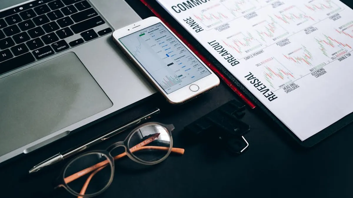 Workstation with laptop, smartphone, eyeglasses, financial charts, and pen for trading analysis.