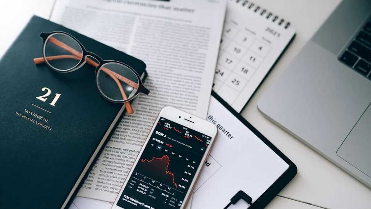 A stylish workspace featuring financial documents, eyeglasses, an iPhone displaying stock data, and a laptop.