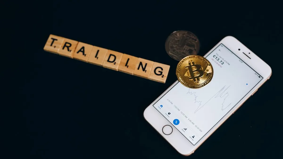 Cryptocurrency trading concept featuring Bitcoin, Scrabble tiles, and a smartphone on a black surface.