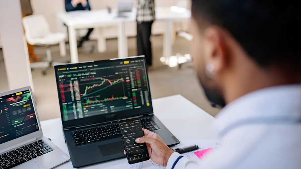 A businessman using smartphone and laptop to track stock market trends in a modern office setting.