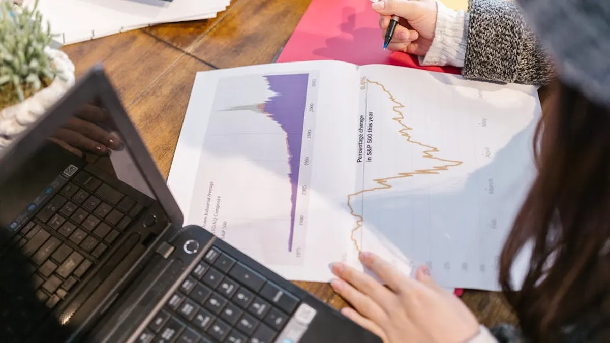 Person reviewing financial graphs and charts while taking notes beside a laptop.