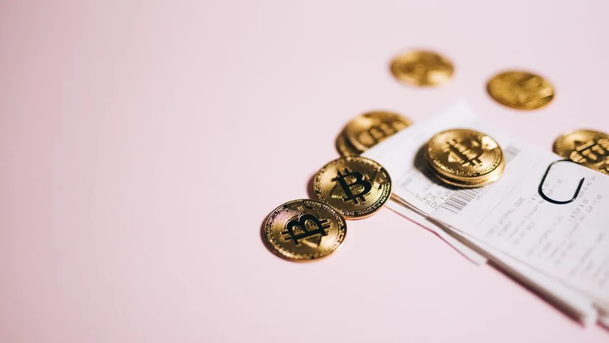 Close-up of Bitcoin coins and cash receipts on a soft pink background.