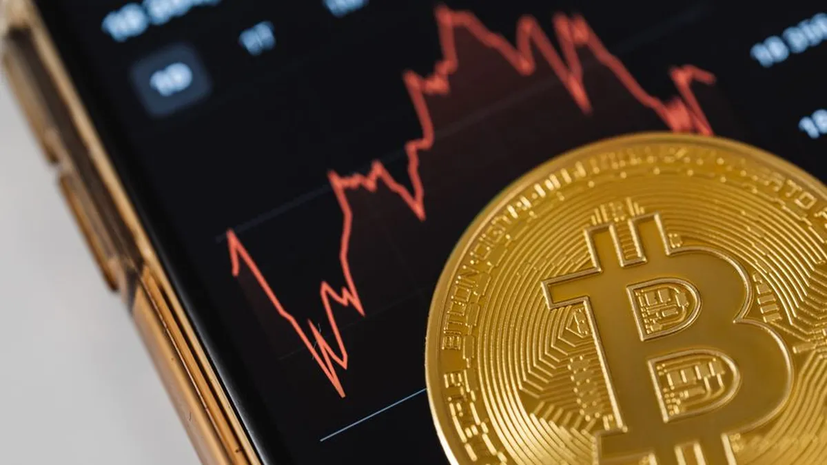 A gold Bitcoin coin on a smartphone displaying a fluctuating stock market chart.