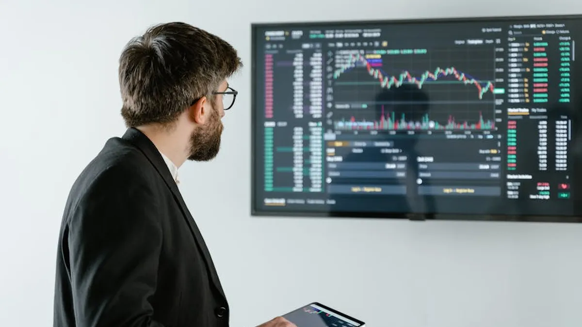 Man observing stock market data on a screen and holding a tablet, representing financial analysis and trading.