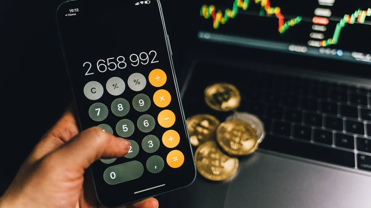 A smartphone displays a calculator app beside bitcoins and a laptop with a financial chart.